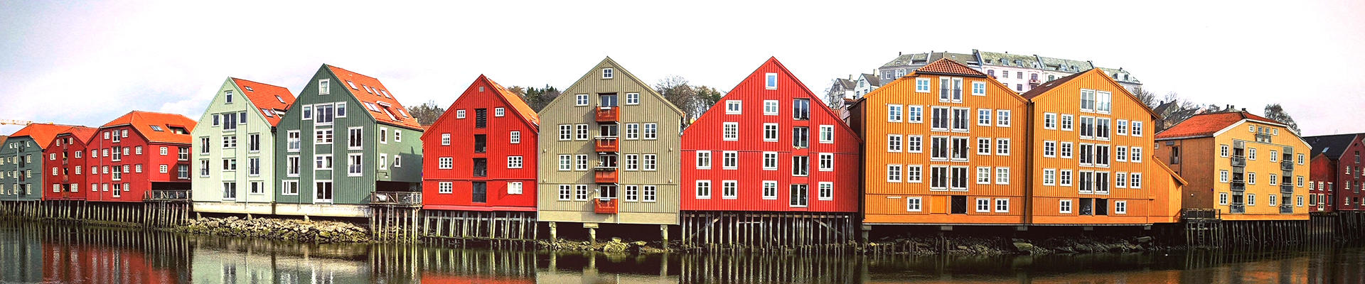 Panoramabilde av en bryggerekke med store bygninger med sterke farger i grønt, rødt, hvitt, grått og gult. Bryggene står langs en elv.